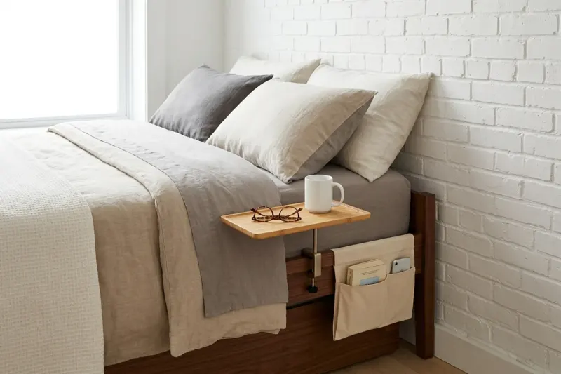 AI concept illustration of a compact rental bedroom featuring a bamboo clamp-on bed-rail tray and a canvas mattress pocket in place of a nightstand.